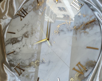 May include: A round, white marble clock face with gold Roman numerals and gold hands. The clock face has a subtle gold marbling effect.