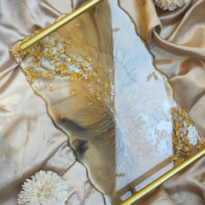 Könnte beinhalten: Ein goldfarbenes und weißes Serviertablett aus Harz mit einem goldenen Metallgriff. Das Tablett hat eine einzigartige, unregelmäßige Form und ist mit Blattgold verziert.