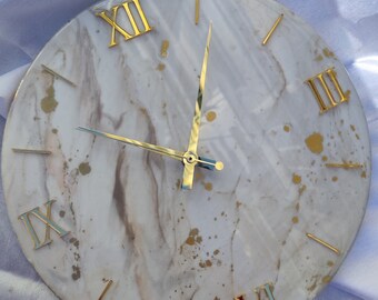 May include: A white and gold clock with Roman numeral markings. The clock face has a marble-like design with gold accents.
