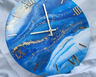 Puede incluir: Reloj de pared redondo con un diseño veteado azul y blanco, acentuado con números romanos y manecillas doradas. La esfera del reloj tiene un patrón abstracto y fluido, creando una pieza decorativa y artística para la decoración del hogar.
