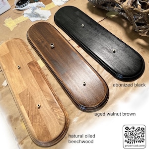 Op de afbeelding: Drie houten displayplaten in natuurlijk geolied beukenhout, verouderd walnootbruin en ebbenzwart. Elke plaat heeft een messingkleurig montagegat. De platen zijn ovaal van vorm met afgeronde randen.