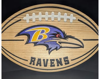 May include: A wooden football-shaped wall sign featuring the Baltimore Ravens logo in purple, gold, black, and white. The design includes a white lace detail at the top and the word RAVENS in black lettering at the bottom, all carved into light wood.
