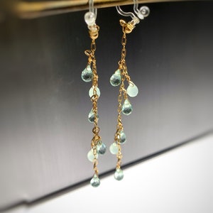 May include: A pair of gold dangle earrings with light green teardrop-shaped gemstones. The earrings are attached to a clip-on earring back.