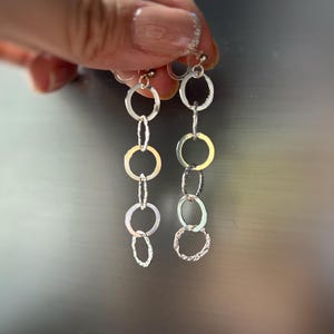 May include: Silver dangle earrings featuring a series of interconnected circles. The earrings have a simple, elegant design with varying textures on the circles. The earrings are held up by a clear earring back.
