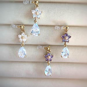 Può includere: Orecchini pendenti color oro con design floreali e a goccia. Gli orecchini presentano piccoli fiori scintillanti bianchi e viola, abbinati a pietre preziose chiare a forma di pera. Gli orecchini sono esposti su uno sfondo neutro.