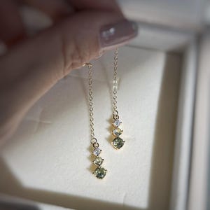 Peut inclure: Une paire de boucles d'oreilles pendantes dorées. Chaque boucle d'oreille est ornée d'une fine chaîne et de trois pierres précieuses : une claire, une vert clair et une autre claire. Les boucles d'oreilles sont présentées sur fond blanc.