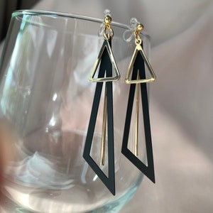 以下が含まれることがあります： 金色のアクセントが付いた黒い三角形のイヤリングのペア。イヤリングは幾何学的なデザインで、透明なプラスチックのポストから吊り下げられています。