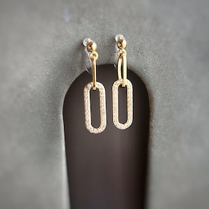 May include: A pair of gold-colored drop earrings. Each earring features a small, round stud at the top, connected to a vertical bar, which leads to a rectangular, open-link design encrusted with small, clear stones. The earrings are displayed against a gray background.