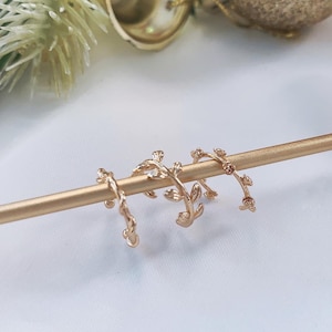 Pode incluir: Três brincos de folha de ouro em um suporte de exibição dourado. Os brincos são delicados e têm um design floral.
