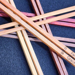 Könnte beinhalten: Holzstäbchen in verschiedenen Braun- und Rottönen, kreuzweise angeordnet. Ein Paar ist mit den Worten "Faith" und "Grace" graviert. Die Stäbchen liegen auf einer dunkelblauen, strukturierten Oberfläche.