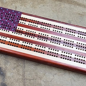 Op de afbeelding: Een houten cribbage-bord met een Amerikaans vlagontwerp. Het bord is gemaakt van verschillende soorten hout, waaronder esdoorn, walnoot en kers. Het bord heeft een paarse en witte sterrenbanner in de linkerbovenhoek, en de rest van het bord bestaat uit rode en witte strepen. Het bord heeft gaten voor pinnen, en de nummers 1 tot en met 12 zijn op het bord gedrukt.