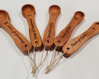 Peut inclure: Ensemble de cinq cuillères à mesurer en bois avec une poignée en ficelle. Chaque cuillère est gravée d'une mesure différente et d'une phrase fantaisiste : "Mound of Love", "Heap of Happiness", "Sprinkle of Joy", "Dash of Glee", et "Pinch of Glory".
