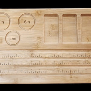 Puede incluir: Una bandeja de rodadura de madera con tres hendiduras circulares etiquetadas como 7 pulgadas, 6 pulgadas y 5 pulgadas. La bandeja también tiene cuatro compartimentos rectangulares y una regla en la parte inferior.