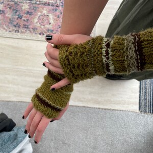May include: Hand-knitted fingerless gloves in shades of green, brown, and cream. The gloves feature a textured pattern and striped accents. These warm, cozy gloves are perfect for cool weather and add a touch of handmade charm to any outfit.