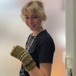 May include: A knitted fingerless glove in shades of green, brown, and cream. The glove features a textured pattern with horizontal stripes and a diamond design. The person is wearing a black t-shirt and a pearl necklace.