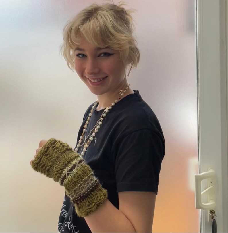 May include: A knitted fingerless glove in shades of green, brown, and cream. The glove features a textured pattern with horizontal stripes and a diamond design. The person is wearing a black t-shirt and a pearl necklace.