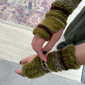 May include: Hand-knitted fingerless gloves in shades of olive green, brown, and cream. The textured knit design features ribbed cuffs and striped accents. These warm wrist warmers are perfect for cool weather.