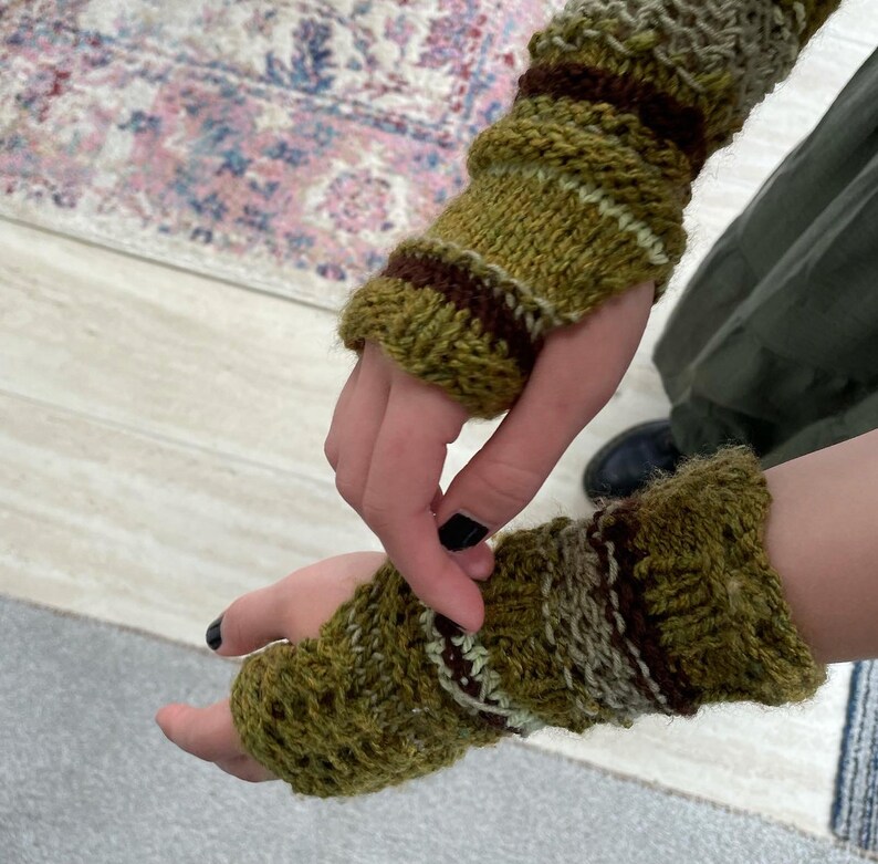 May include: Hand-knitted fingerless gloves in shades of olive green, brown, and cream. The textured knit design features ribbed cuffs and striped accents. These warm wrist warmers are perfect for cool weather.