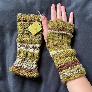 May include: A pair of olive green and brown knitted fingerless gloves. The gloves feature a textured design with white and brown accents. A small tag with a floral design hangs from one glove.