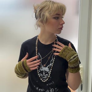May include: A black t-shirt with a graphic design, paired with a long pearl necklace and layered gold necklaces. The outfit includes green and brown knitted fingerless gloves. The person is wearing a black hair clip.