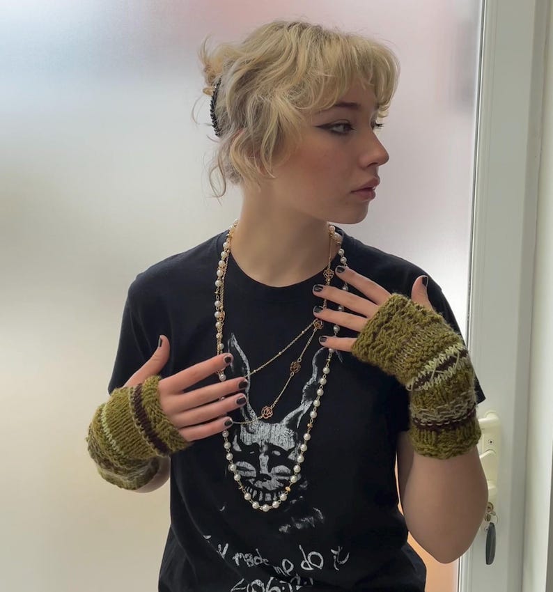 May include: A black t-shirt with a graphic design, paired with a long pearl necklace and layered gold necklaces. The outfit includes green and brown knitted fingerless gloves. The person is wearing a black hair clip.