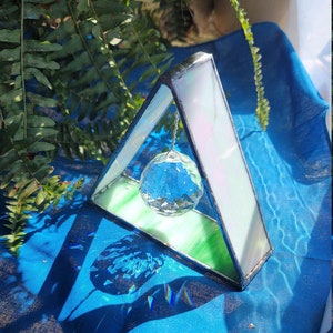 May include: A stained glass triangle with a crystal ball hanging inside. The triangle is made of clear, white, and green glass. The crystal ball is reflecting light and casting rainbows on the blue fabric below.