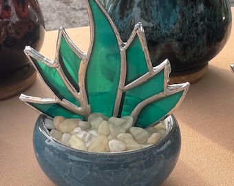 May include: A decorative glass succulent sculpture with emerald green leaves and silver edging, set in a small, round, blue ceramic pot filled with small, light-colored pebbles. The sculpture is a unique home decor item.