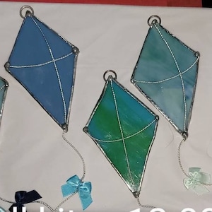 May include: Six stained glass kites in various shades of blue and green. The kites are hanging from a string and have a white background.