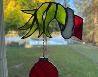 May include: A stained glass ornament featuring the Grinch's hand holding a red ornament. The Grinch's hand is green and yellow, and the ornament is red with white stripes.