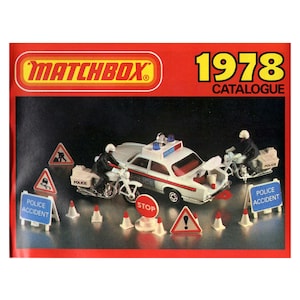 Puede incluir: Una página del catálogo Matchbox de 1978 que presenta un coche de policía blanco con una luz azul en la parte superior, dos motocicletas de policía y varias señales de tráfico y conos. El texto "Matchbox" y "Catálogo 1978" es visible en la parte superior de la página.