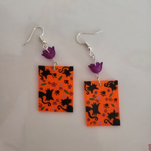 May include: A pair of orange and black cat earrings with purple flower accents. The earrings are rectangular and feature a repeating pattern of black cats, paw prints, and flowers on an orange background.