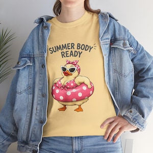 Peut inclure: Un t-shirt jaune clair avec une illustration de canard portant un bikini à pois roses, un bandeau assorti et des lunettes de soleil, dans une bouée rose à pois. Le texte au-dessus du canard indique SUMMER BODY READY.