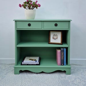 May include: A painted sage green bookcase with two drawers and open shelves. It displays books, a framed owl picture, and a plant with pink flowers. A framed landscape painting hangs above the bookcase.