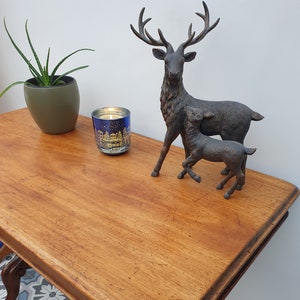 May include: A wooden table with a carved base and a distressed finish. A candle with a winter scene design and two bronze deer figurines are on the table.