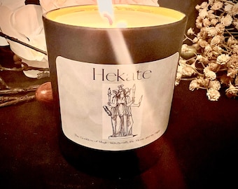 May include: A black candle with a white label that reads "Hekate, The Goddess of Magic, Witchcraft, the Moon and the Night". The candle is lit and the flame is casting a warm glow.