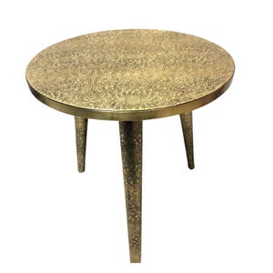 May include: A round, gold-colored metal side table with an intricate, engraved design on the top and three legs.