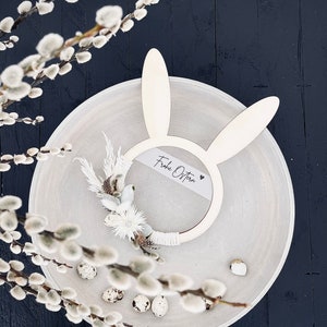 Könnte beinhalten: Ein weißer Hasen-Reifen mit dem Text "Frohe Ostern" und einem kleinen Herz auf einem weißen Teller mit Wachteleiern und Weidenzweigen.