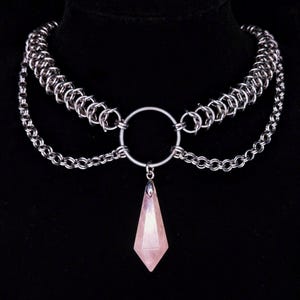 Könnte beinhalten: Eine silberne Ketten-Choker-Halskette mit einem großen, runden Ring und einem rosafarbenen Kristall-Anhänger. Der Anhänger ist in Form eines langen, spitzen Tropfens geformt.