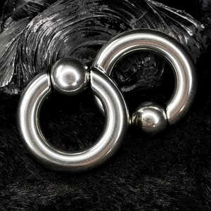 May include: Two silver circular body jewellery pieces with a ball on each side. The jewellery is on a black background.