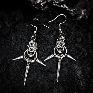 May include: A pair of silver earrings with a gothic design. Each earring has a cross-shaped element with three spikes and a decorative chain. The earrings hang from a simple hook. The background is dark.