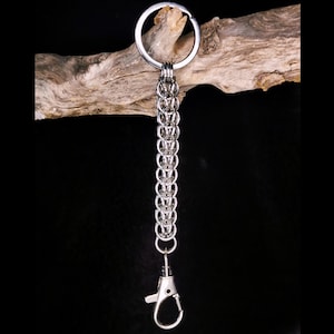 Peut inclure: Un porte-clés argenté avec un design de style cotte de mailles. Le porte-clés comprend un grand anneau en haut, une série d'anneaux métalliques interconnectés formant la chaîne et un mousqueton en bas. Le fond est noir.