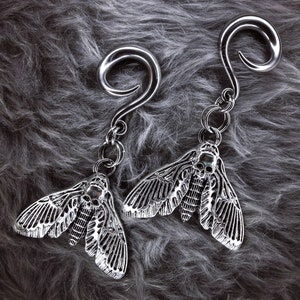 May include: Two silver moth-shaped earrings with skull details hanging from silver hoops. The earrings are on a gray fuzzy surface.