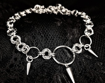 May include: A silver chain choker necklace with three silver spikes and a large silver ring. The necklace is on a black lace background.