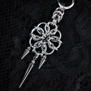 Peut inclure: Pendentif en métal argenté avec un motif floral composé de maillons de type cotte de mailles et trois pointes coniques pendantes. Il est attaché à un porte-clés. Le fond est un tissu noir texturé.