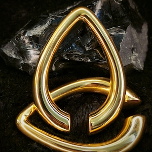 May include: Gold-toned metal body jewellery in the shape of a teardrop with a curved, open design. The jewellery is on a black furry surface.