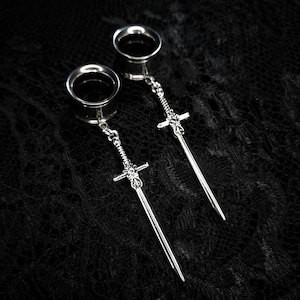 May include: A pair of silver ear gauges with dangling sword charms. The gauges have a circular design, and the swords are detailed with intricate hilt designs. The background is a dark, textured fabric.