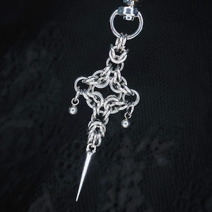 May include: A silver-toned metal chainmail pendant featuring an intricate woven link pattern. The design includes a central cross-like structure with two small spherical beads hanging from the sides and a long, pointed metal spike at the bottom.
