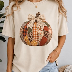 May include: A cream-coloured short-sleeve t-shirt featuring a large central graphic of a pumpkin. The pumpkin is composed of various patchwork fabric patterns including plaid, floral, leopard print, and corduroy, topped with a beige lace bow.