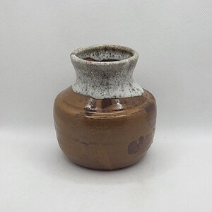 Puede incluir: Un pequeño jarrón de cerámica marrón con un esmalte blanco en la parte superior. El jarrón tiene un cuerpo redondeado y una abertura ligeramente acampanada.