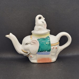May include: A white ceramic teapot shaped like an elephant with a smaller elephant on top of its head. The teapot has a green, blue, yellow, and orange design.